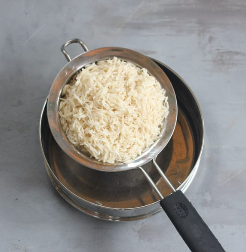 Cooked rice is being drained in a metal strainer placed over a steel bowl on a gray surface.