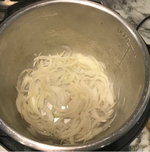 Thinly sliced onions in an Instant Pot on a marble countertop, starting to cook and appear slightly translucent.