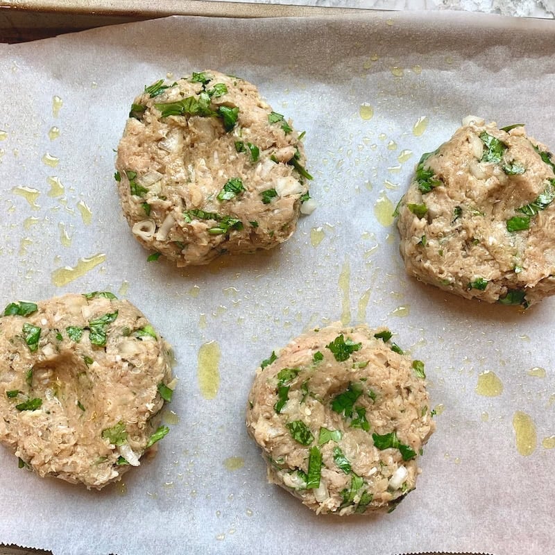 Four uncooked chicken burger patties with chopped herbs on parchment paper, drizzled with oil, ready to bake.