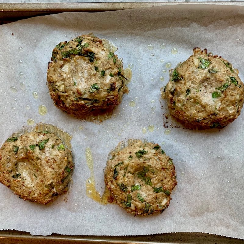 Four baked chicken burger patties on a parchment-lined baking sheet, drizzled with oil.