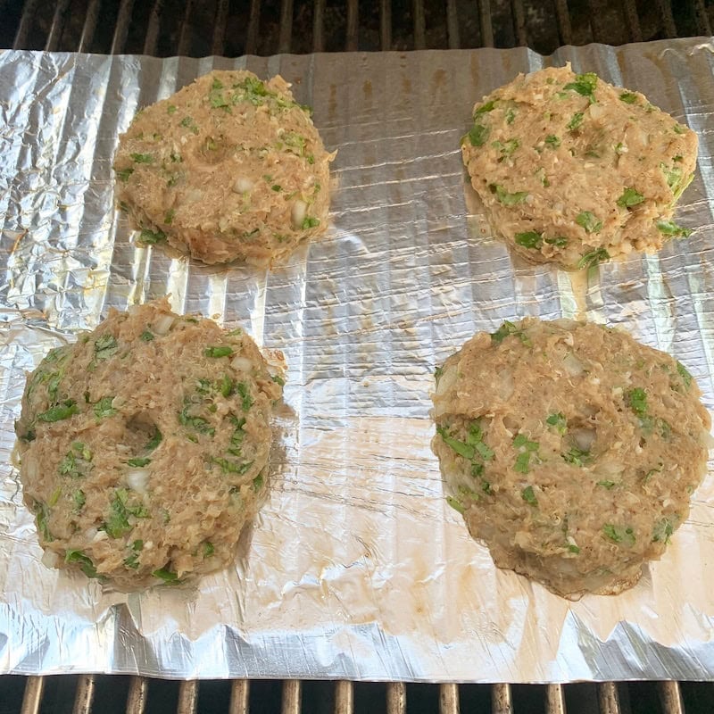 Four uncooked burger patties with visible green herbs sit on a sheet of aluminum foil on a grill.