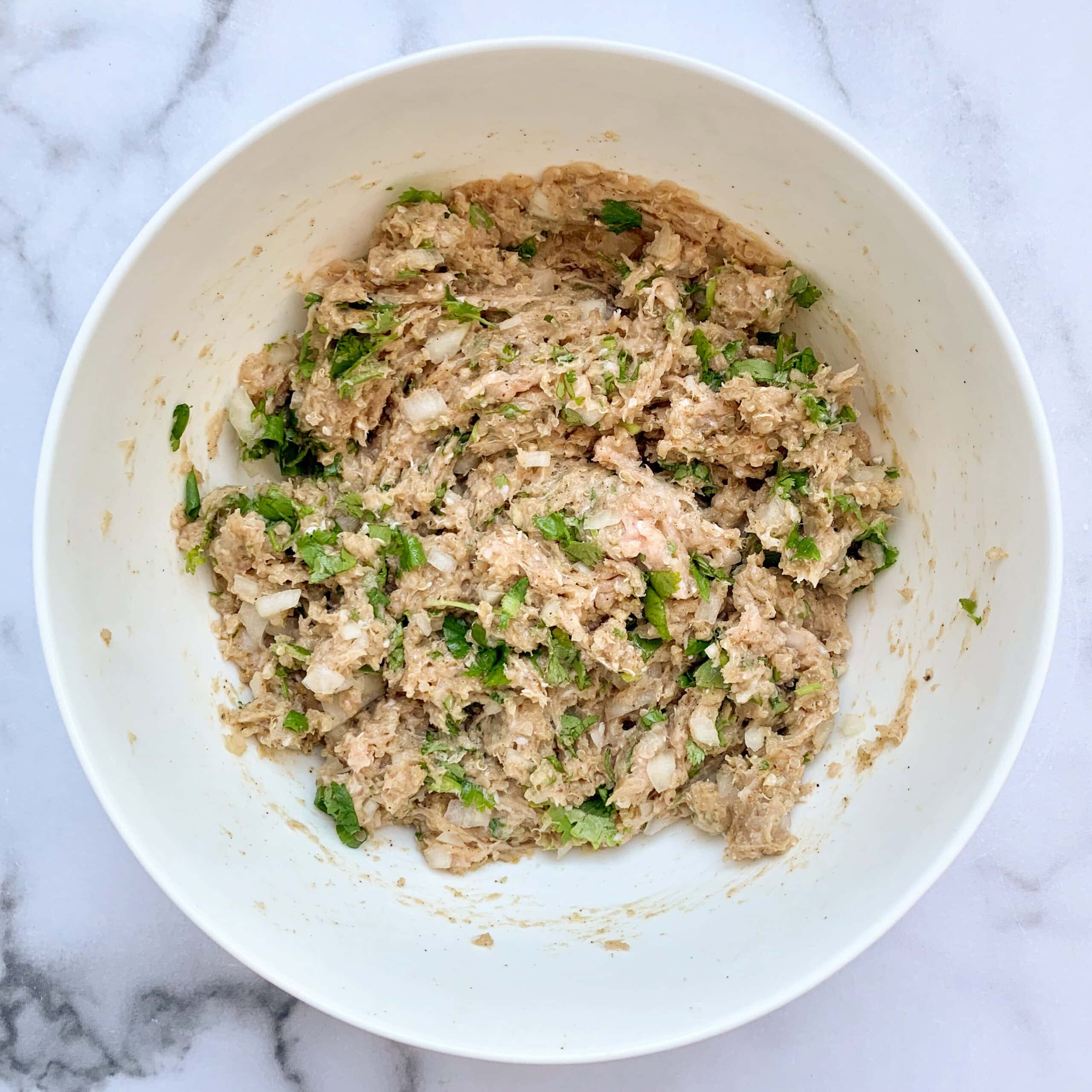A white bowl filled with a mixture of minced meat, chopped onions, and green herbs on a marble surface.