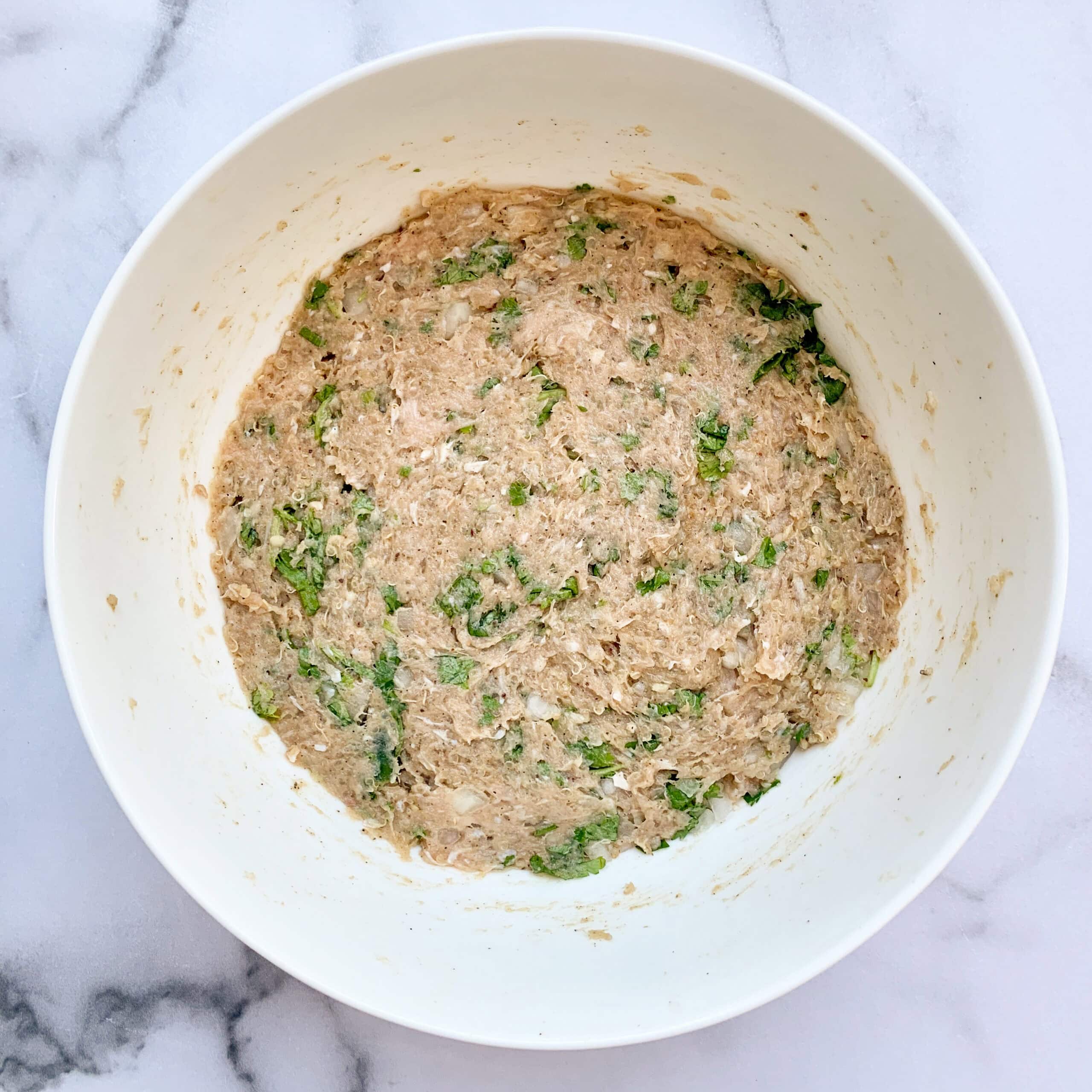 A white bowl containing a mixture of ground meat, chopped herbs, and other ingredients sits on a marble surface.
