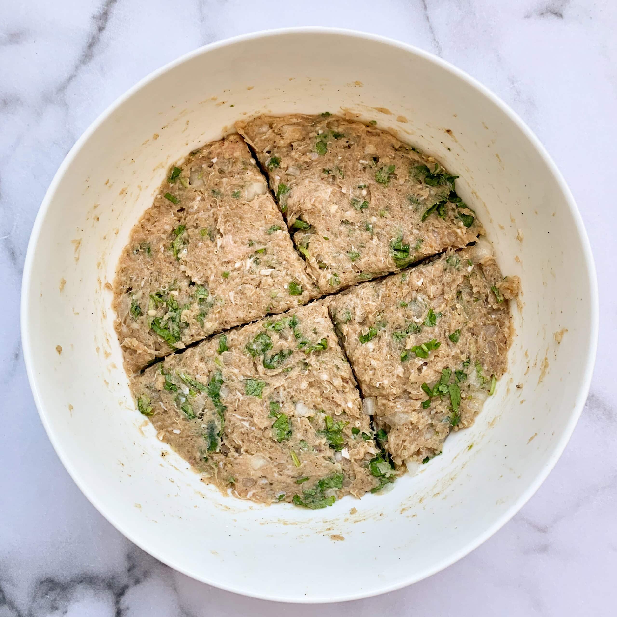 A white bowl containing a mixture of ground meat, herbs, and seasonings, divided into four sections on a marble surface.