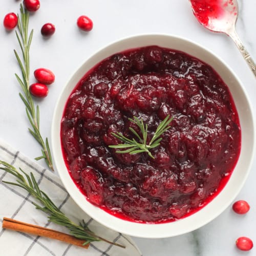 Cranberry sauce in a white bowl with rosemary and cinnamon on the side