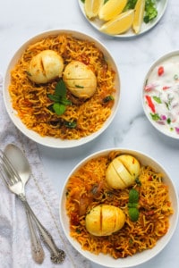 egg biryani served in 2 white bowls with raita and lemon