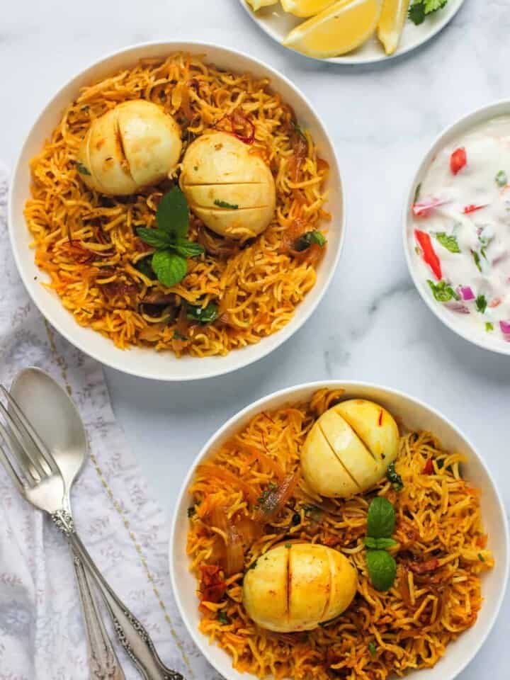 egg biryani served in 2 white bowls with raita and lemon