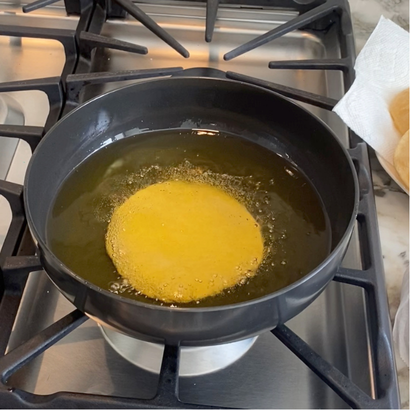 Puri is being fried in hot oil in a black pan on a stovetop, with bubbling visible around the edges.