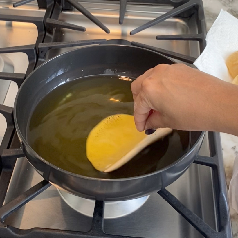 A hand lowers a rolled puri into hot oil in a black pan on a stovetop, preparing to fry it.