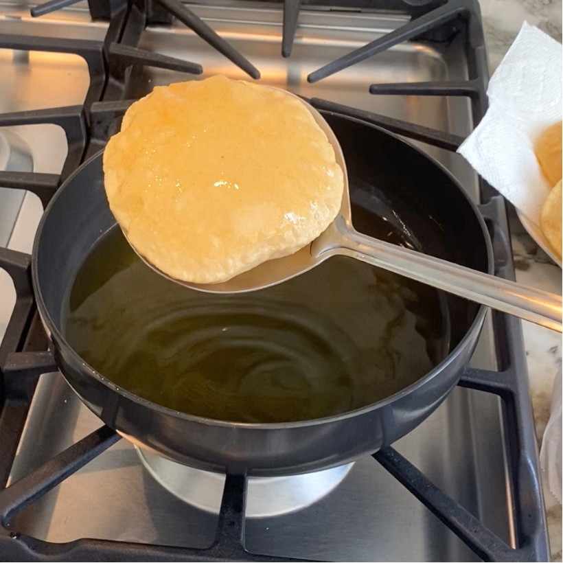 A puffed puri is lifted from hot oil with a slotted spoon over a stovetop, with more puris on a paper towel nearby.