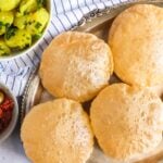 Four puffed, fried puris on a silver tray, with a side of spiced potatoes and pickles placed nearby on a striped cloth.