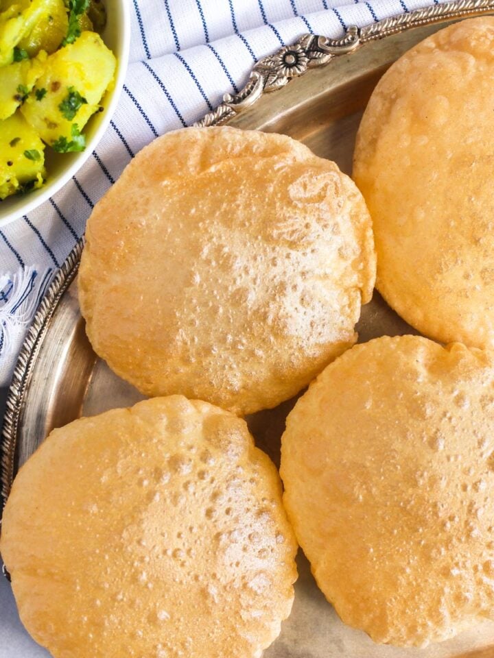 Four puffed, fried puris on a silver tray, with a side of spiced potatoes and pickles placed nearby on a striped cloth.