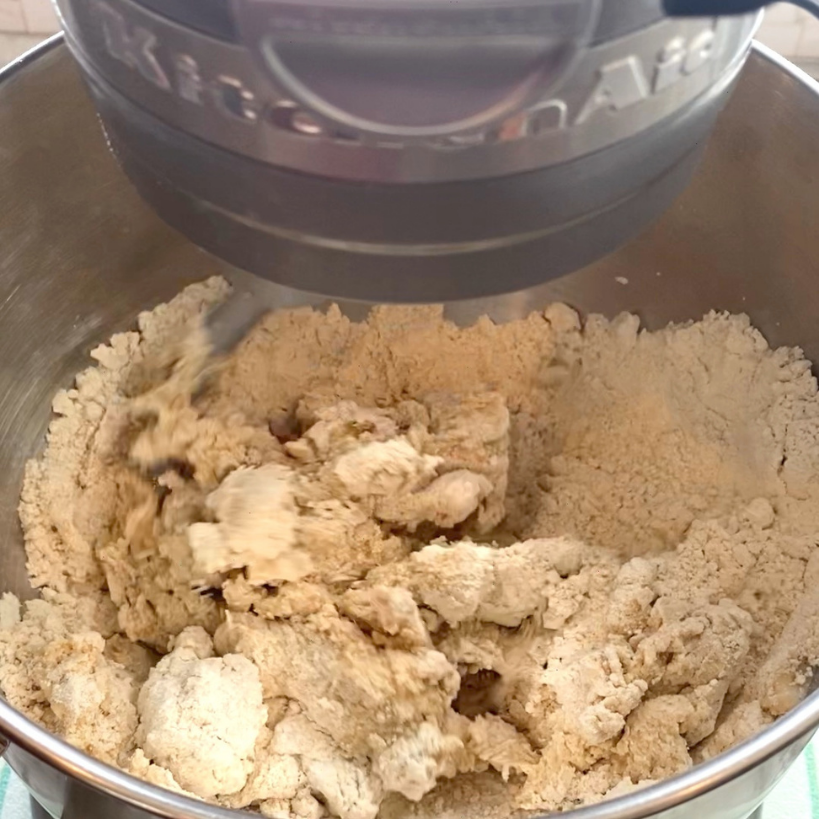 Stand mixer blending dough in a stainless steel bowl, with paddle attachment combining dry and wet ingredients into a rough, crumbly mixture.