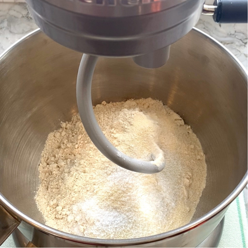 A stand mixer bowl contains a mixture of flour and sugar with a dough hook attachment positioned above the ingredients.