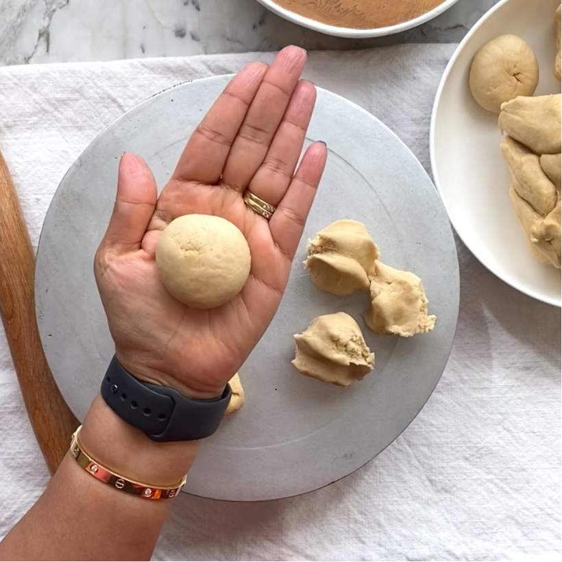 A hand holds a round ball of dough above a flat surface, with more dough balls and pieces visible nearby on a white plate and cloth.