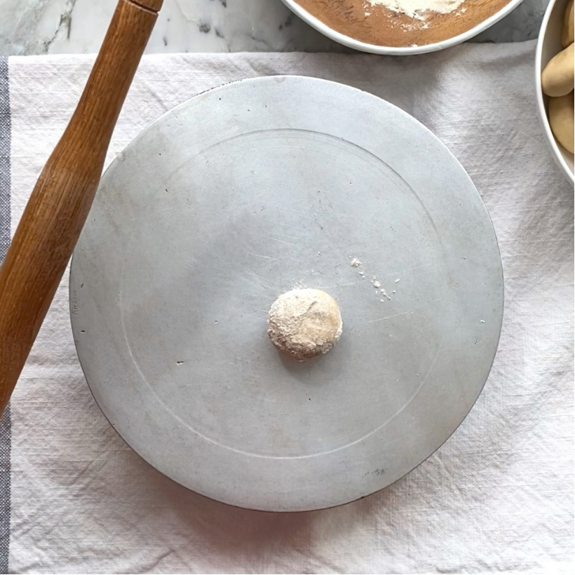 A single dough ball dusted with flour sits on a round metal plate, with a rolling pin and bowls of dough and flour nearby on a cloth-covered surface.