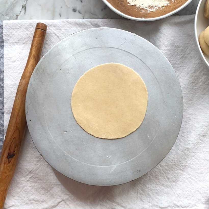 A round, uncooked dough disc sits on a flat gray surface next to a wooden rolling pin and a bowl on a white cloth.