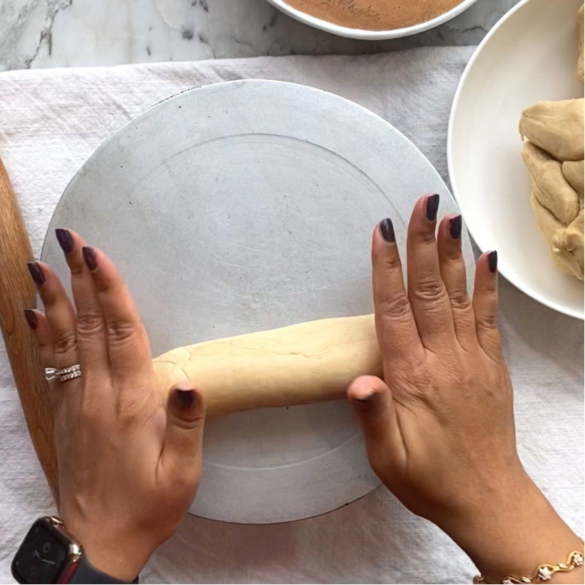 Hands rolling a piece of dough on a round silver surface, with more dough and a bowl visible nearby on a white cloth background.