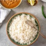 A bowl of cooked basmati rice with a fork, a curry dish, lime wedges, a green chili, and a napkin on a gray surface. Text at the top reads Instant Pot Basmati Rice.