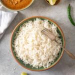 A bowl of cooked white basmati rice with a fork, placed on a gray surface with curry, lime wedges, and a green chili nearby.