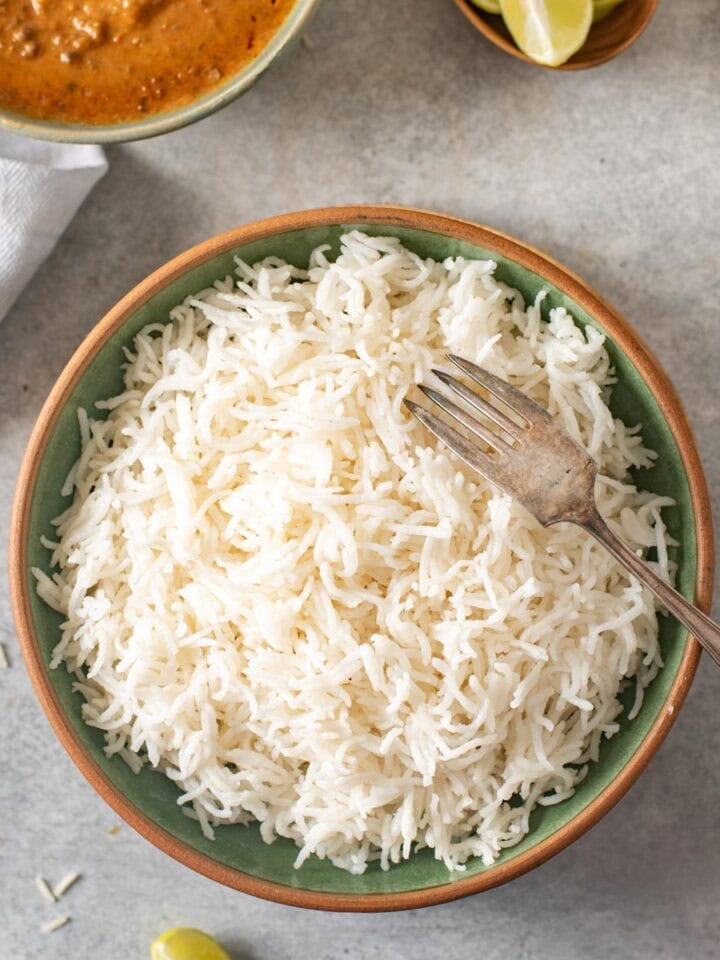 A bowl of cooked white basmati rice with a fork, placed on a gray surface with curry, lime wedges, and a green chili nearby.