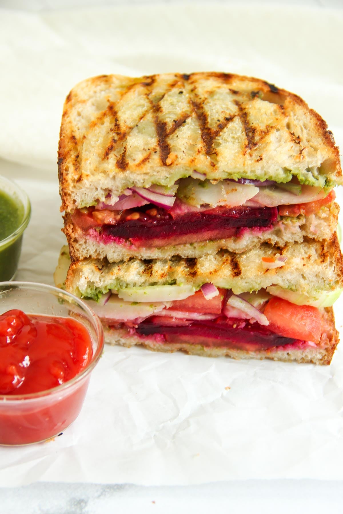 A stacked grilled vegetable sandwich with visible layers of beetroot, tomato, cucumber, and onion, served with small cups of green chutney and red sauce on the side.