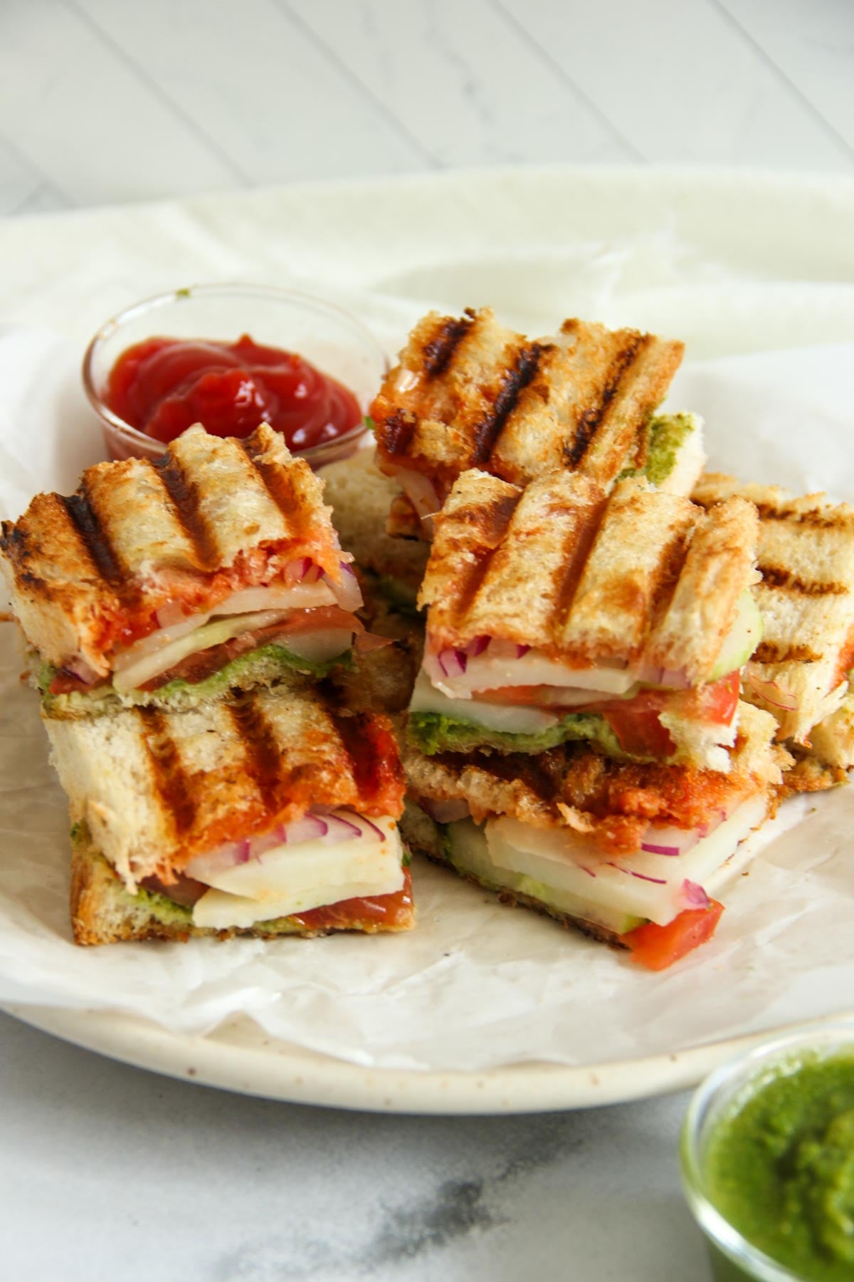 Stacked grilled vegetable and cheese sandwiches on a plate, served with ketchup and green chutney in small bowls.