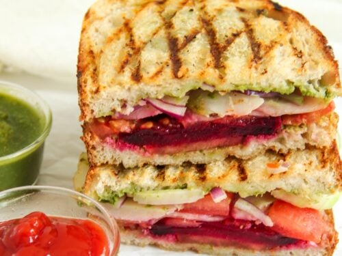 Grilled vegetable sandwich stacked in halves, filled with tomato, beet, cucumber, and onion, served with green chutney and ketchup in small bowls.