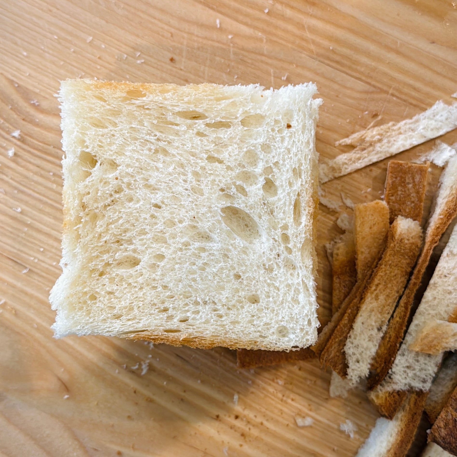 A square slice of white bread with crusts removed sits on a wooden surface next to a pile of bread crust strips.