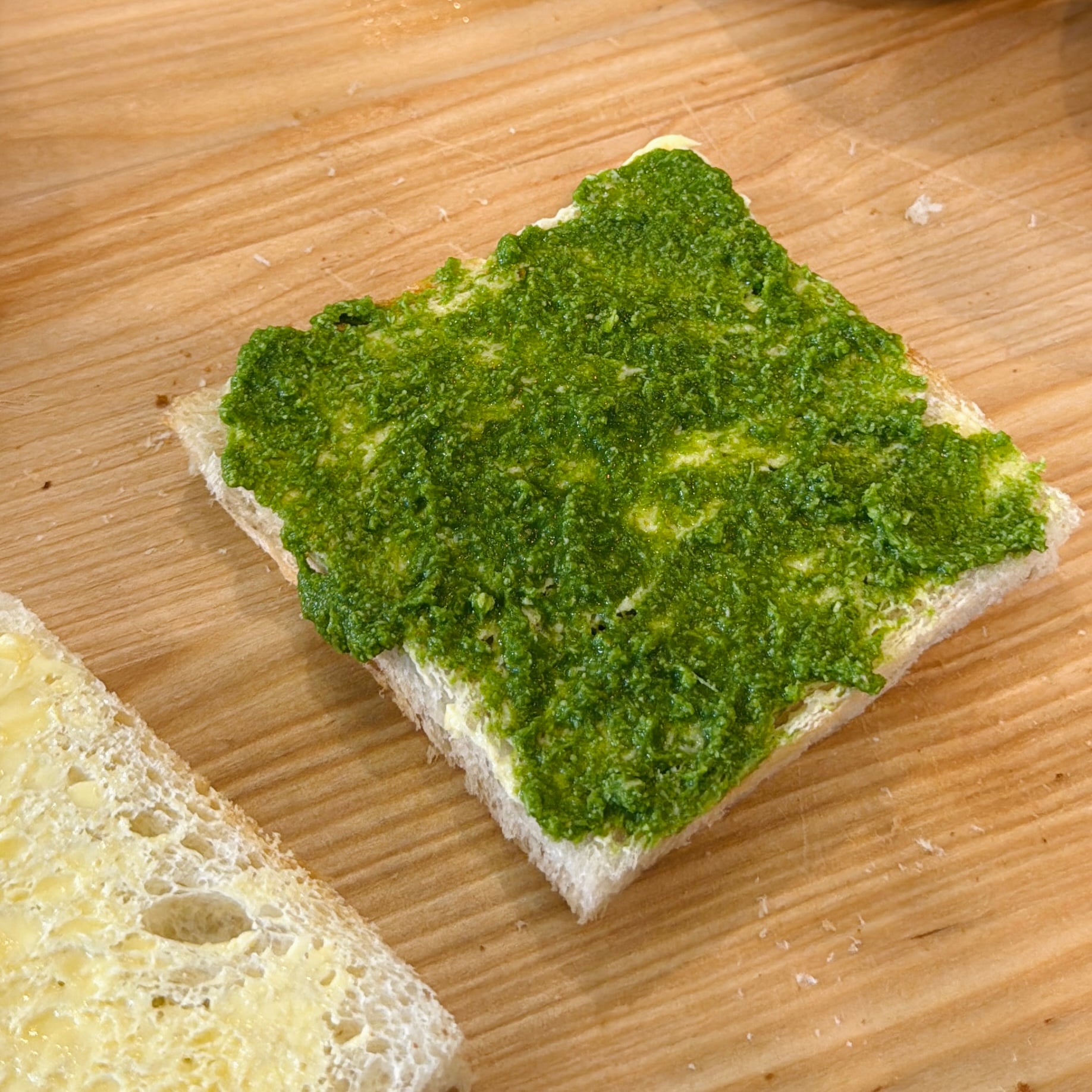 A slice of white bread topped with a green pesto spread sits on a wooden surface next to another slice with a yellow spread.