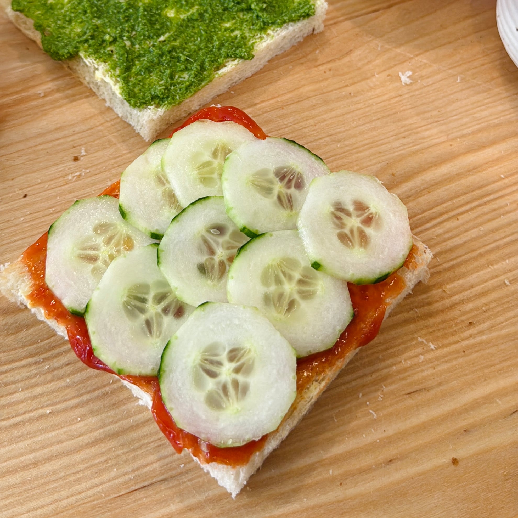 A slice of bread topped with sliced cucumbers and a red spread sits on a wooden surface; another slice with green spread is nearby.