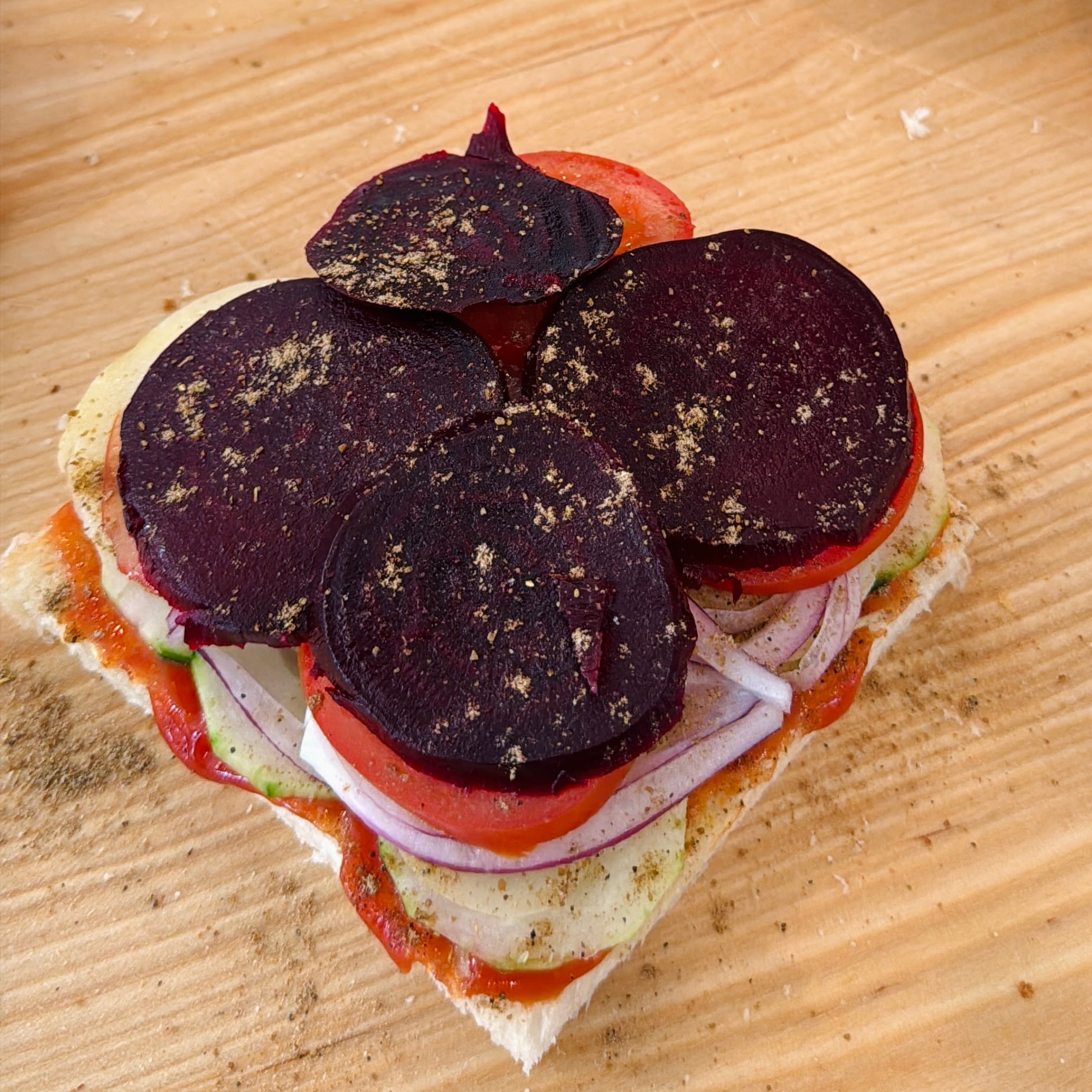 Open sandwich on white bread with layers of sliced beetroot, tomato, onion, and potato, sprinkled with black pepper, on a wooden surface.
