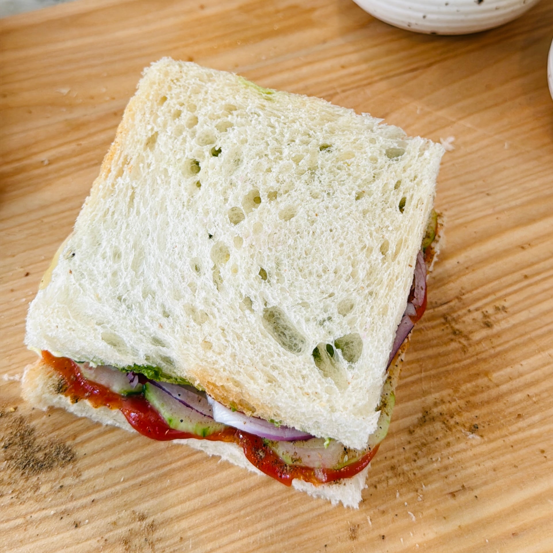 A sandwich with white bread, visible layers of sliced cucumber, onion, green chutney, and ketchup on a wooden surface.