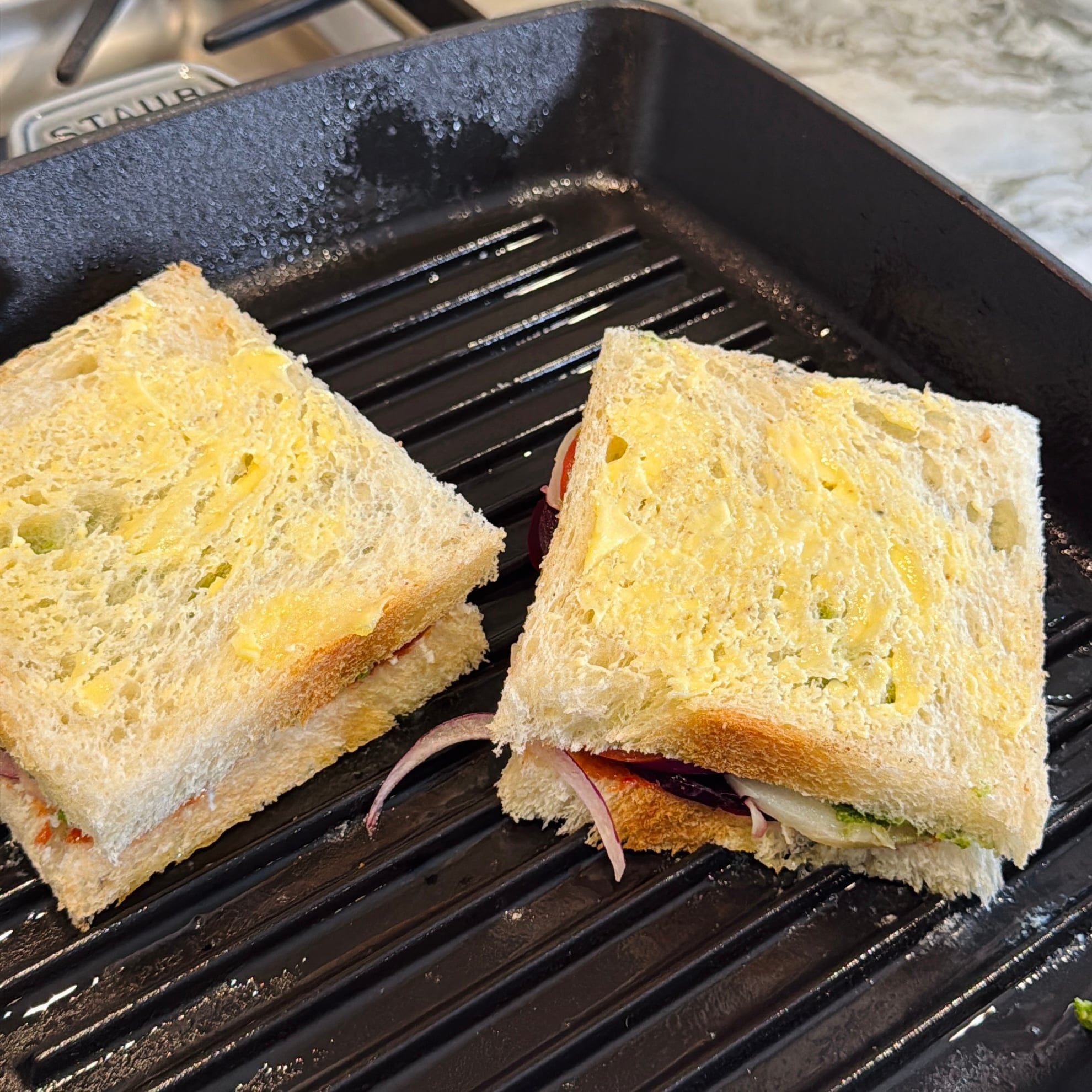 Two buttered sandwiches with visible onion slices are placed on a black stovetop grill pan, ready to be cooked.