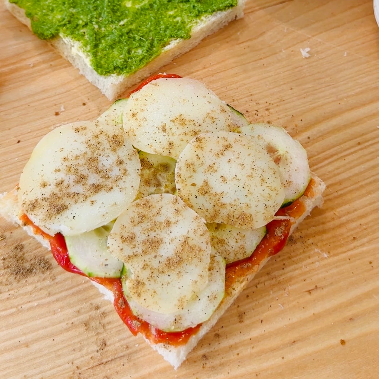 A slice of bread topped with sliced potatoes, cucumbers, tomatoes, and sprinkled spices, next to another slice spread with green chutney, all on a wooden surface.