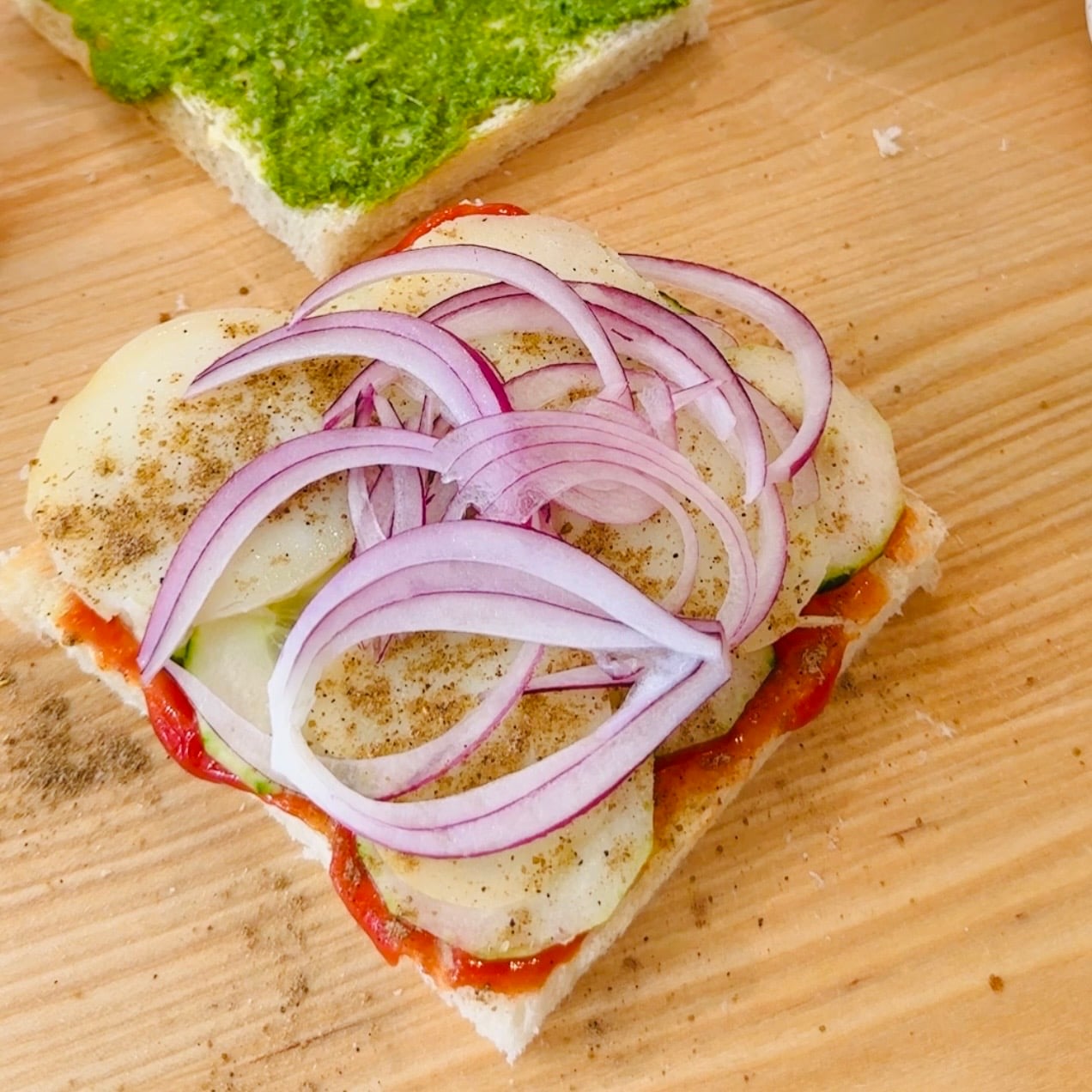 A slice of bread topped with tomato sauce, cucumber, potato slices, red onion, and black pepper. Another bread slice with green spread is nearby on a wooden surface.