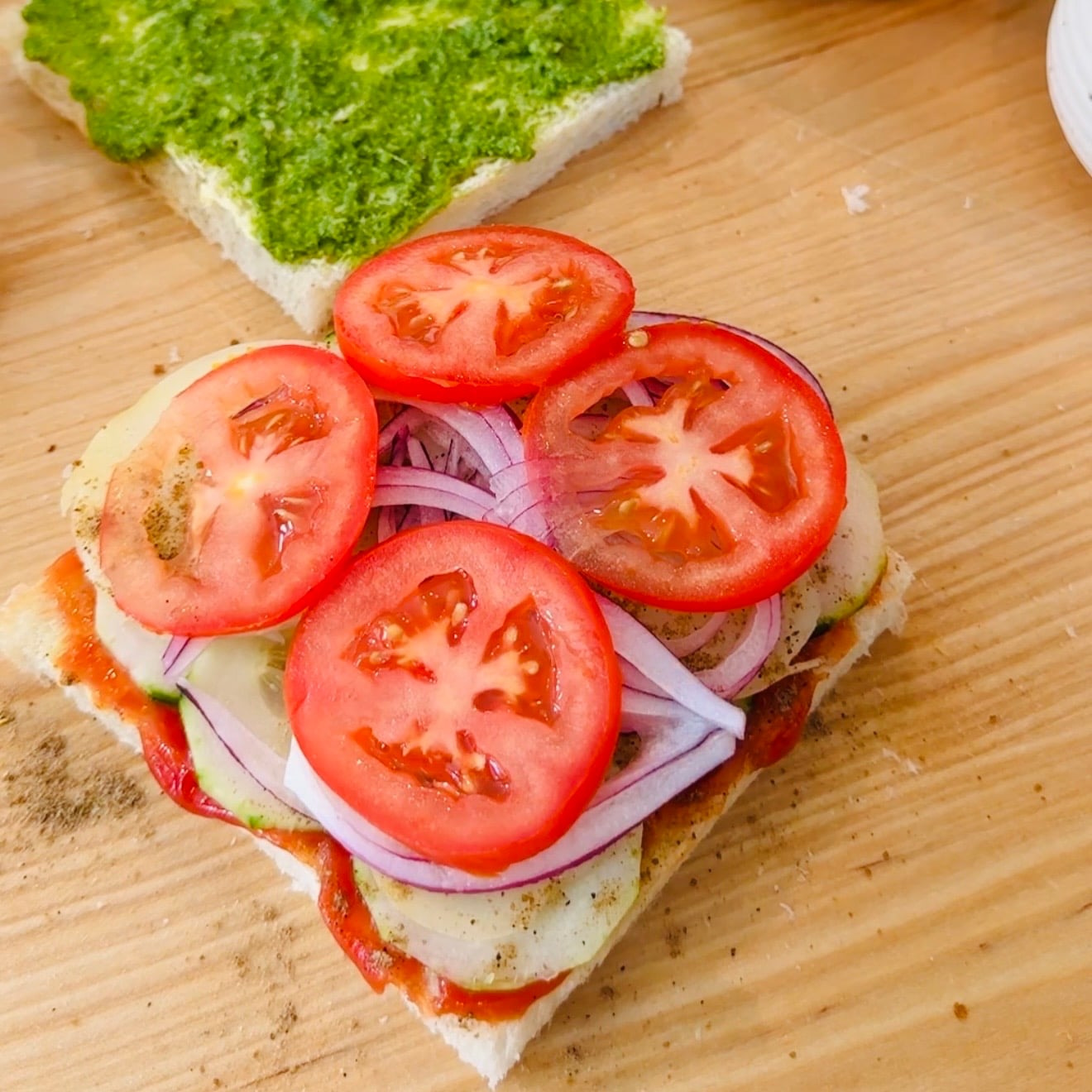 Open sandwich on white bread topped with sliced tomatoes, red onions, cucumbers, and seasoning; another slice with green spread is in the background.