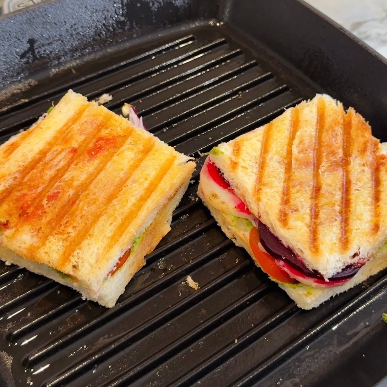 Two vegetable paninis with grill marks sit on a black ridged grill pan, showing layers of sliced vegetables and toasted bread.