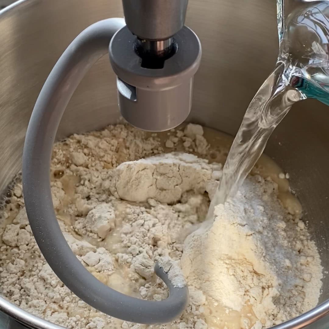 Flour and other dry ingredients in a stand mixer bowl with water being poured in, ready for mixing.