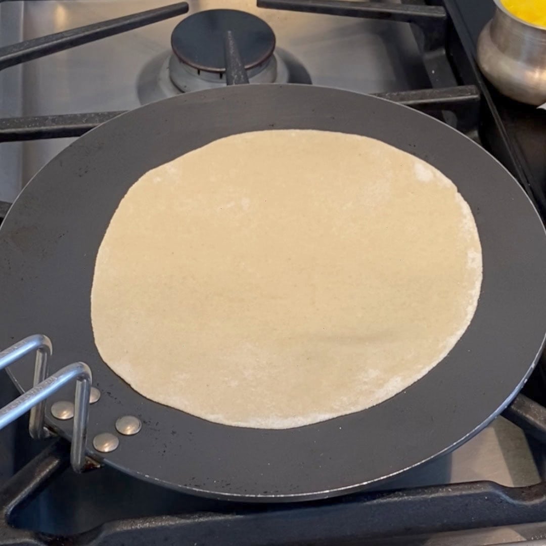 A rolled-out dough circle is cooking on a flat skillet over a stove burner.