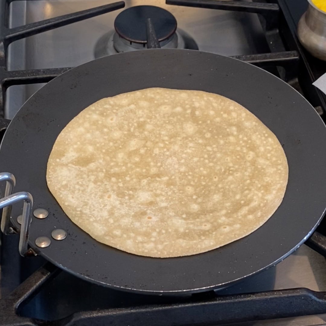 A round flatbread is cooking on a black pan over a gas stove.