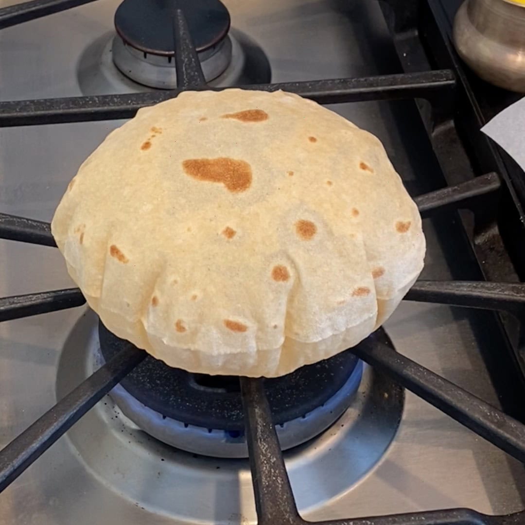 A puffed roti is cooking directly over a gas stove burner on a metal grate.
