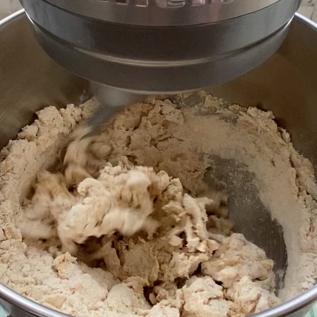 Stand mixer blending flour and dough in a metal bowl, with the paddle attachment in motion.