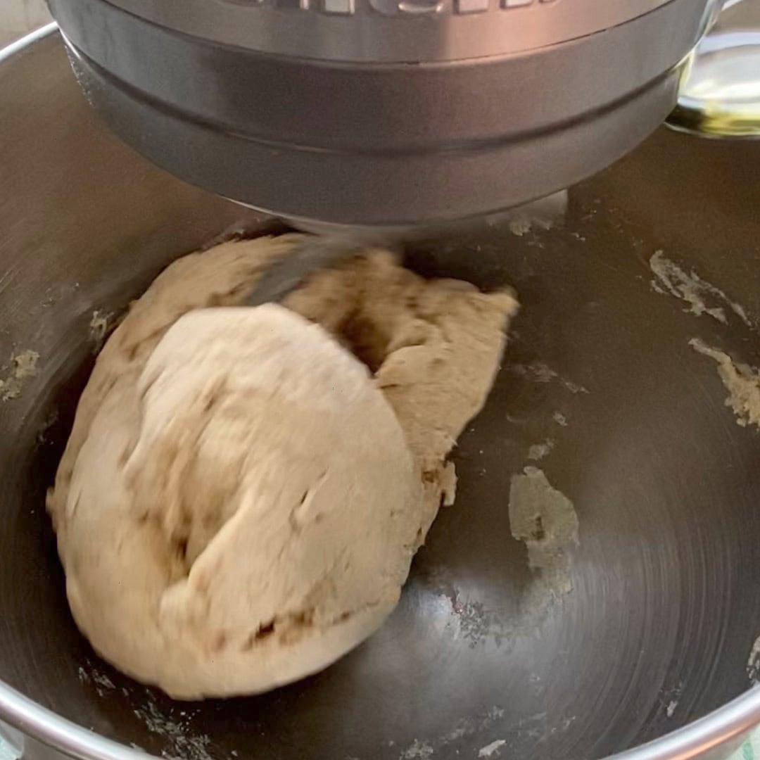 A stand mixer is kneading a ball of bread dough in a metal mixing bowl.
