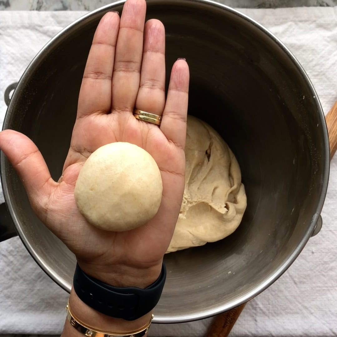 A hand holds a smooth dough ball above a mixing bowl containing more dough, with a white cloth underneath the bowl.
