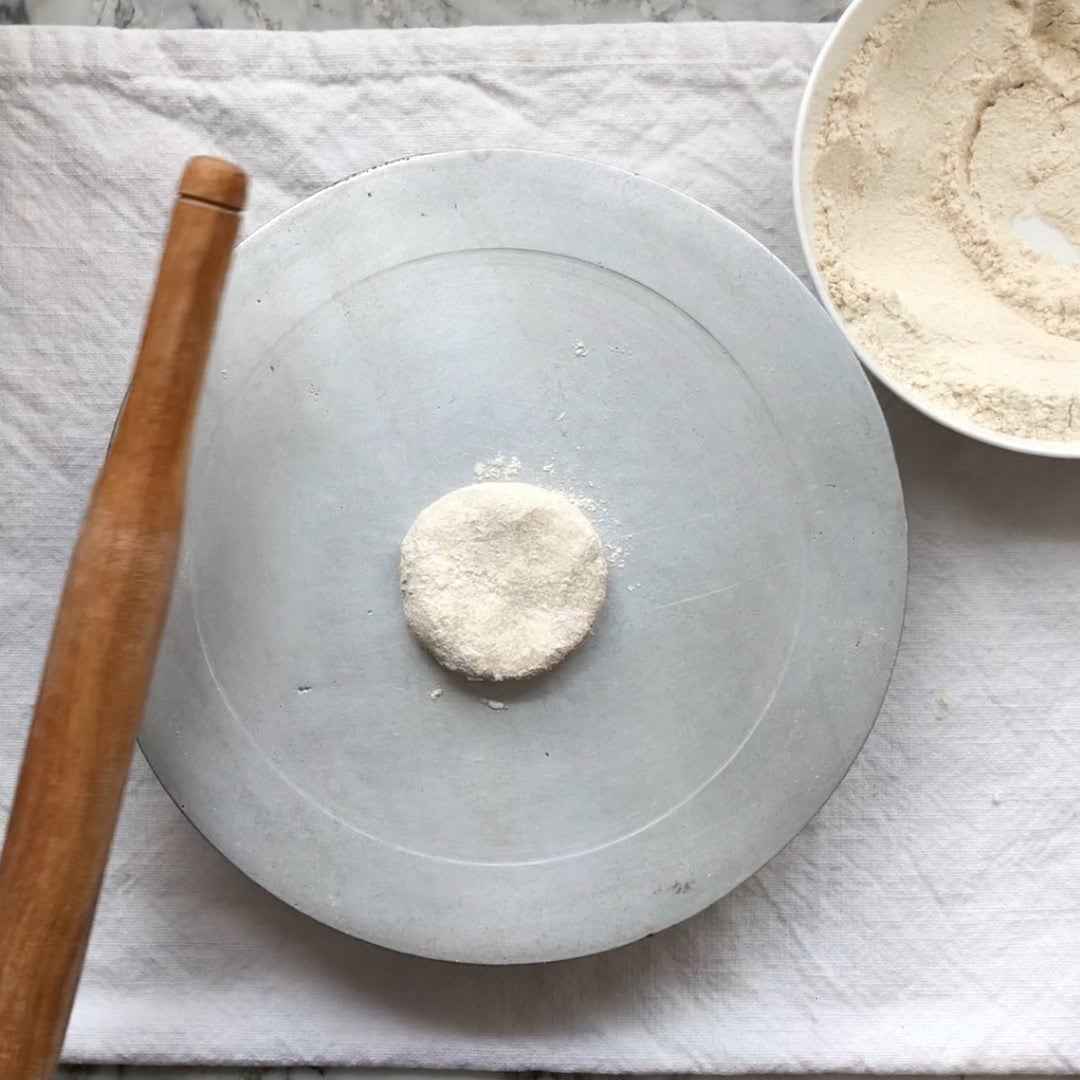 A small ball of dough sits on a round metal board next to a wooden rolling pin and a bowl of flour on a white cloth surface.