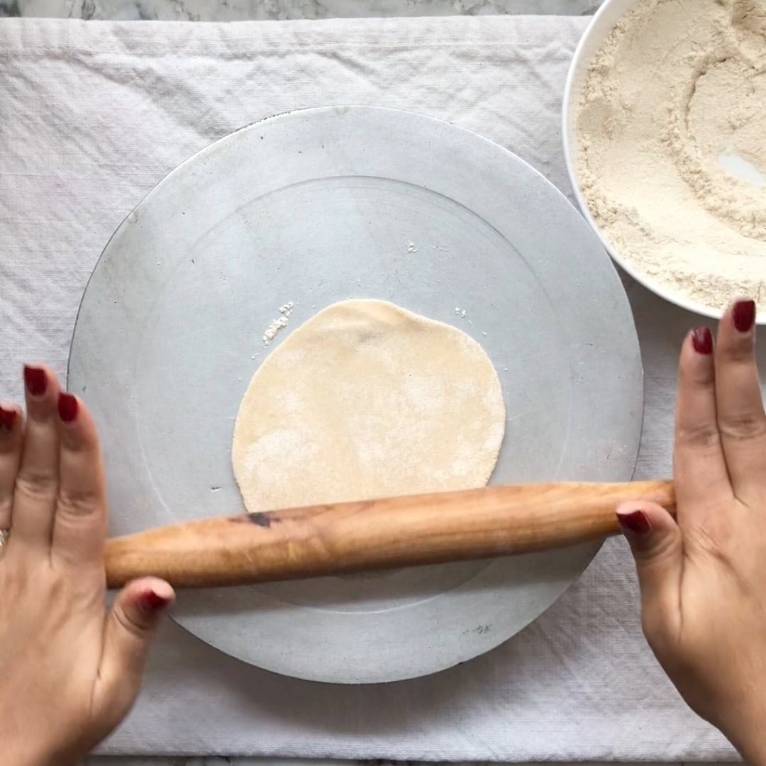 Hands rolling out dough with a rolling pin on a flat surface, with a bowl of flour nearby.