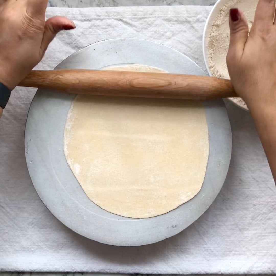 Person rolling out dough into a flat circle on a round board with a wooden rolling pin, next to a bowl of flour.