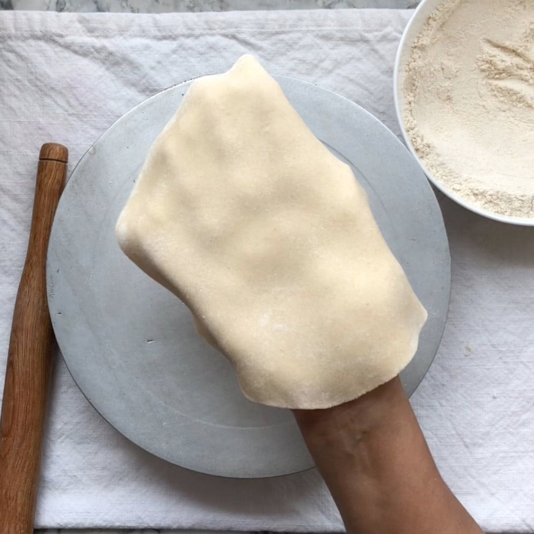 A hand holds a sheet of rolled dough over a metal plate, with a rolling pin and a bowl of flour nearby on a white cloth.
