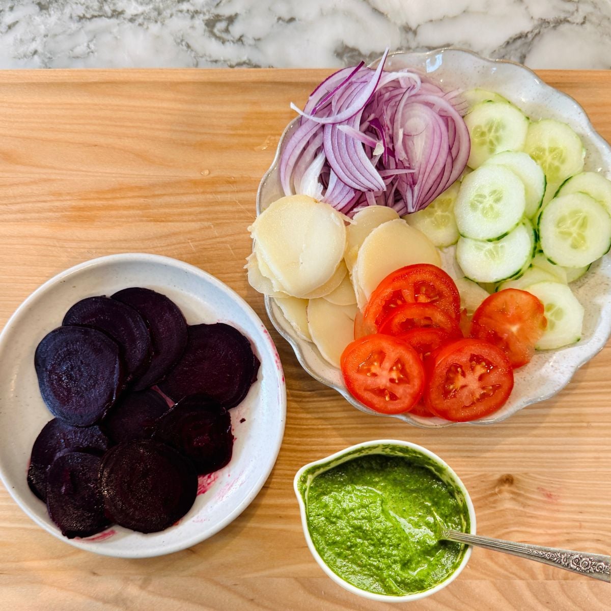 Sliced cucumbers, tomatoes, potatoes, and red onions on a plate, sliced beets in a bowl, and green chutney in a small dish on a wooden surface.